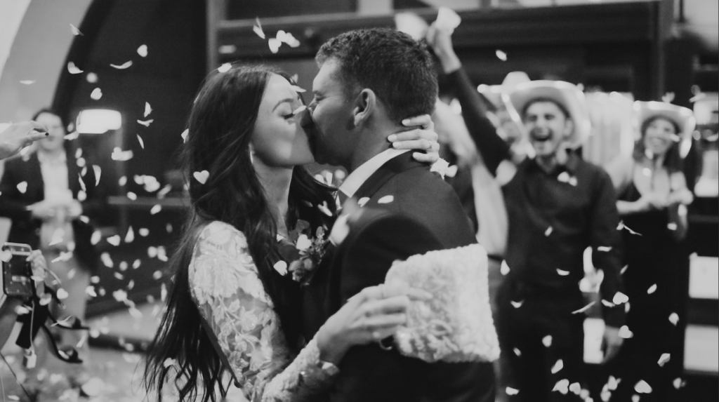 black and white photo of bride and groom kissing under falling white confetti